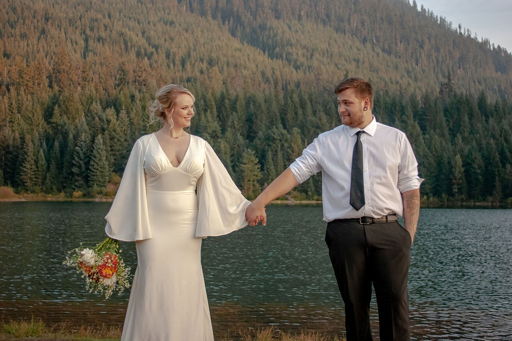 Luxury Elopement Experience in the Pacific North West. Couple Eloping while holding hands with a beautiful lake and mountains full of green trees behind them.