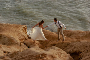 Couple enjoy and budget adventure elopement along side the cliffs of San Diego Beach. Groom is reaching Brides hand